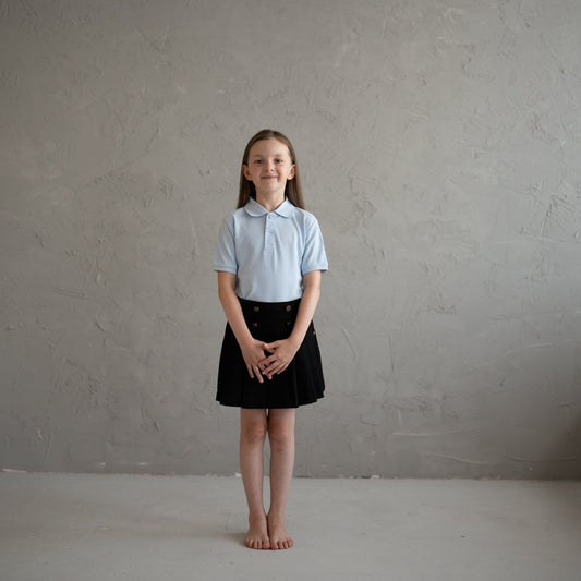 School girl wearing a light blue shirt and black pleated skirt standing against a plain gray background