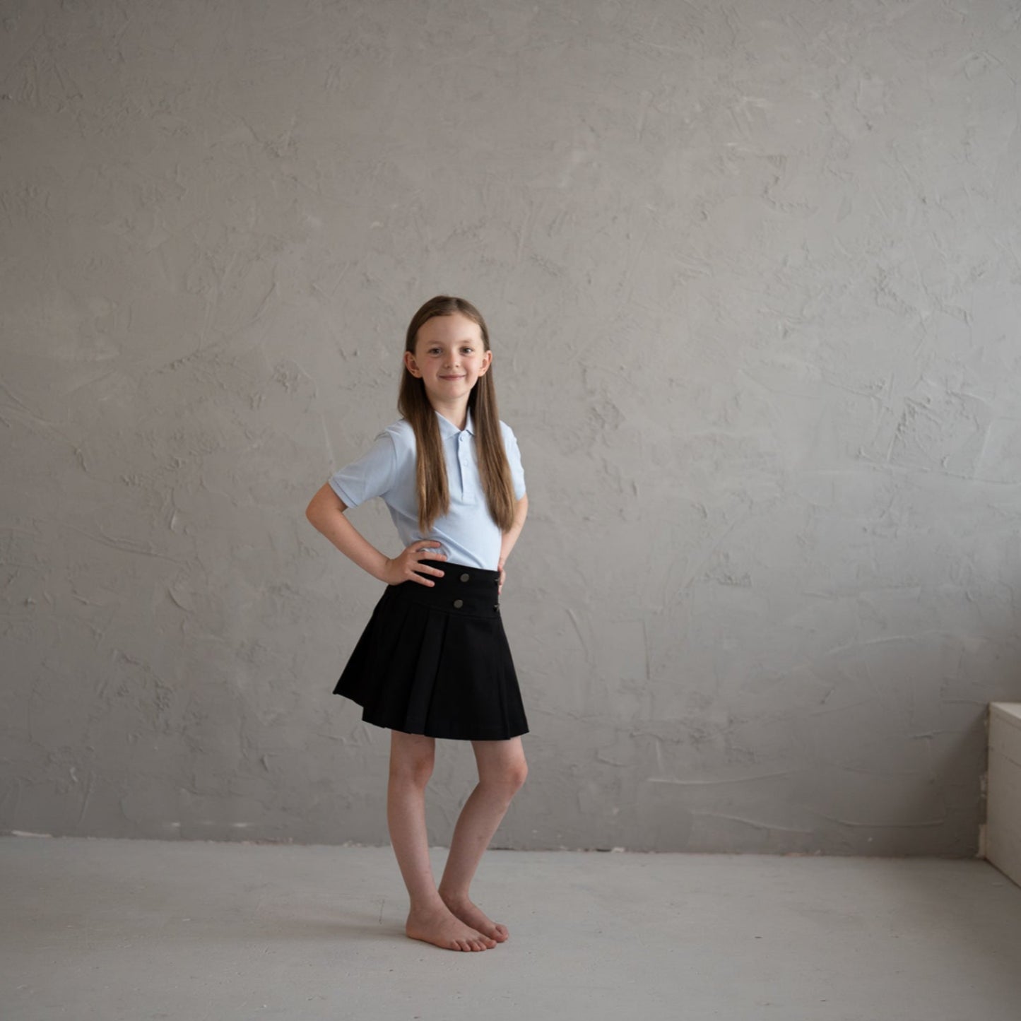Young girl in a white shirt and black skirt standing in an empty room with a gray wall.