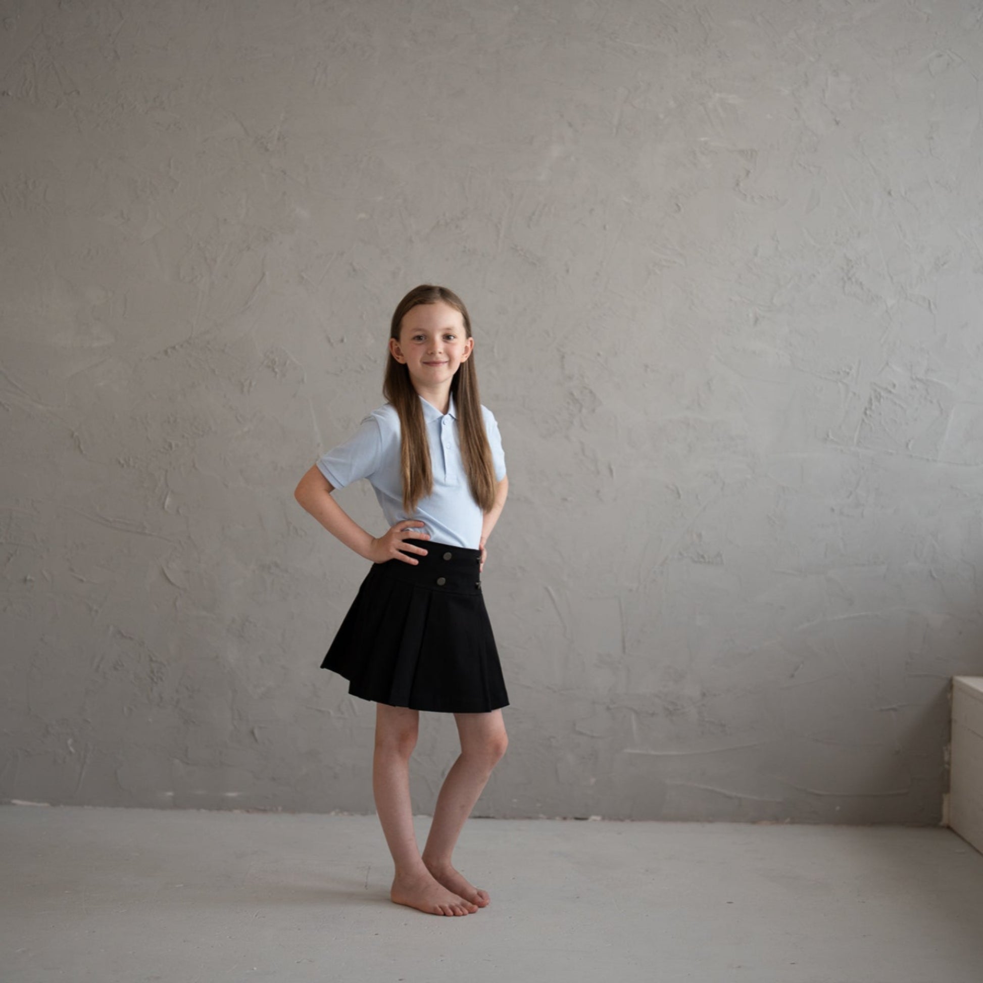 Young girl in a white shirt and black skirt standing in an empty room with a gray wall.